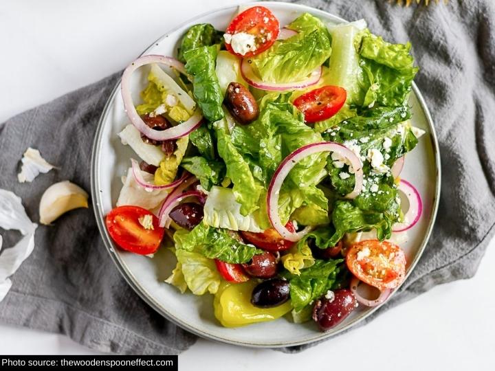 Sliced red onion, tomatoes, olives, and lettuce with feta cheese on a white plate on a grey tablecloth.