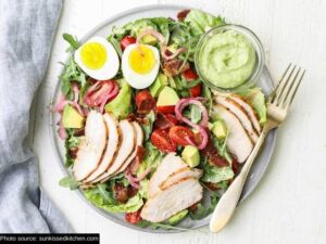 Sliced chicken breast, red onion, avocado, a sliced egg, and lettuce on a plate with a cup of green goddess dressing.