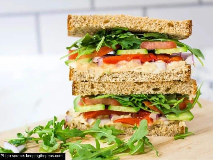 A sandwich sliced in half with the two halves stacked on top of one another. The sandwich has lettuce, tomato, cucumber, red onion, and hummus.