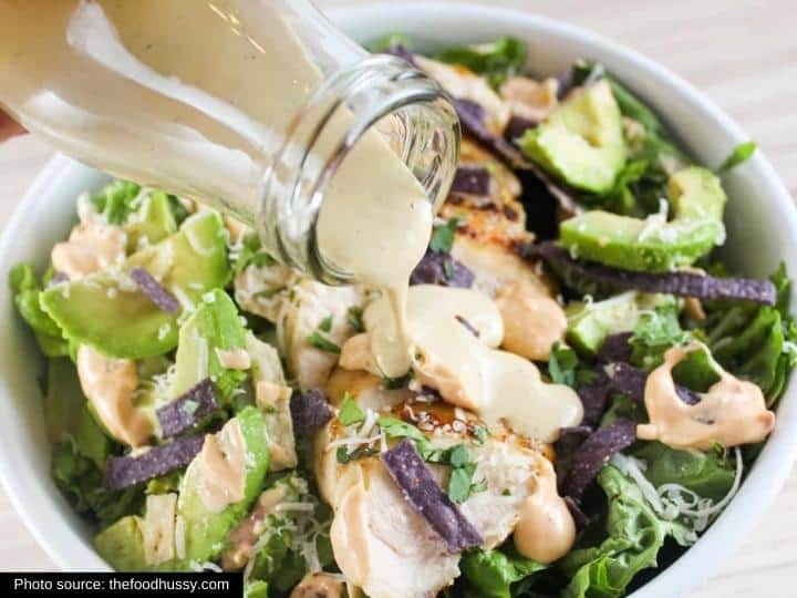 A bottle pouring dressing on top of chicken breasts, avocado, lettuce, and blue tortillas in a white bowl.