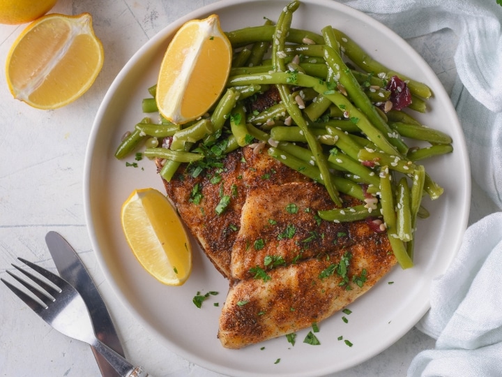 Green beans and a piece of cooked tilapia with two lemon wedges on a plate.