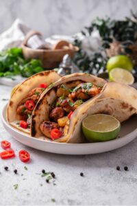 A plate with two chicken al pastor tacos with a halved lime on the side. The tacos contain chicken, pineapple, and peppers with cilantro on top. There is a wooden bowl with seasonings and limes in the background.