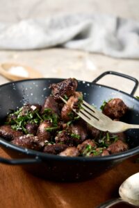 A ceramic bowl filled with cooked chicken hearts topped with fresh parsley. The bowl is on a wooden server and there is a fork taking a bite of chicken hearts.