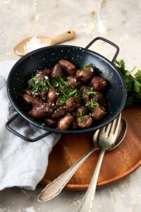 A ceramic bowl filled with cooked chicken hearts topped with fresh parsley. The bowl is on a wooden server with a fork, spoon, and napkin next to the bowl.