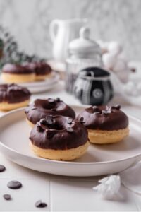 A ceramic plate with three baked donuts with chocolate frosting on top. The donuts are sprinkled with chocolate chips and there is an additional plate of donuts in the background.