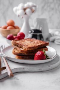 Four slices of french toast stacked on a white plate on top of a serving board. There is a strawberry and fork on the side.