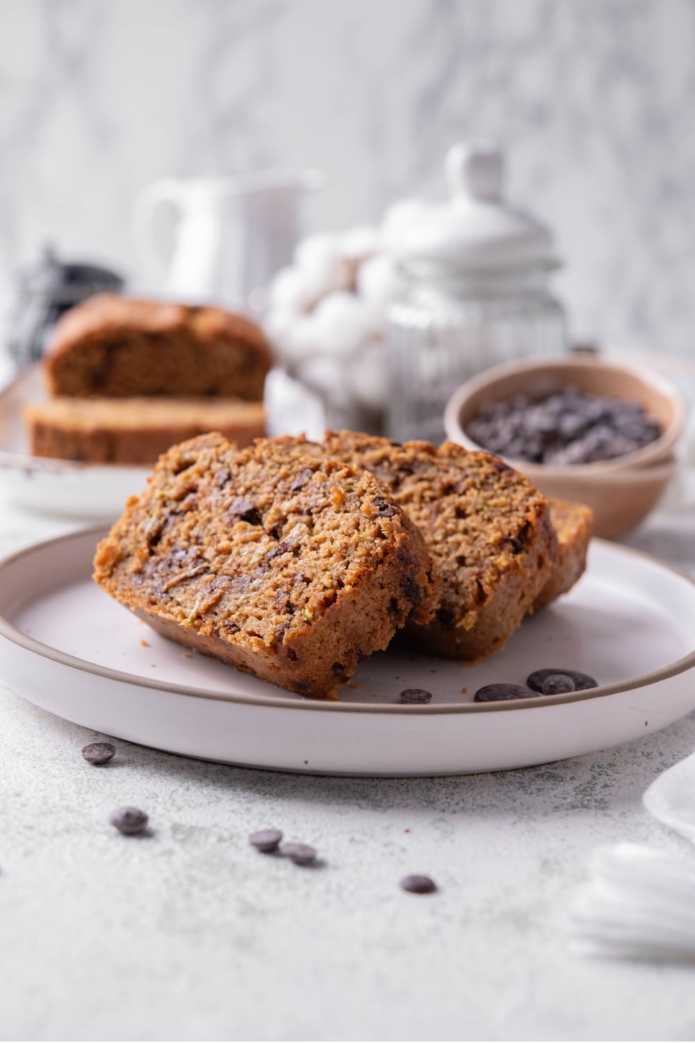 Three slices of low calorie zucchini bread with chocolate chips arranged on a white ceramic plate. There are a few chocolate chips scattered with another loaf of bread and bowl of chocolate chips in the background.