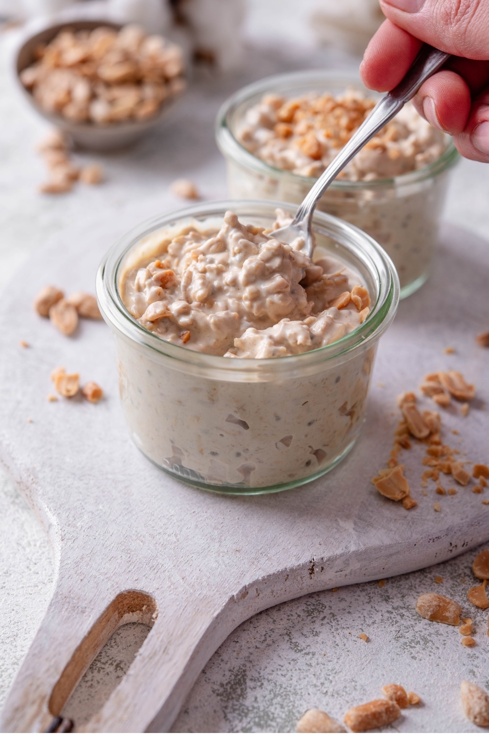 A small glass jar filled with peanut butter overnight oats on top of a wooden serving board with peanuts sprinkled around it. There is a hand dipping a spoon into the jar.