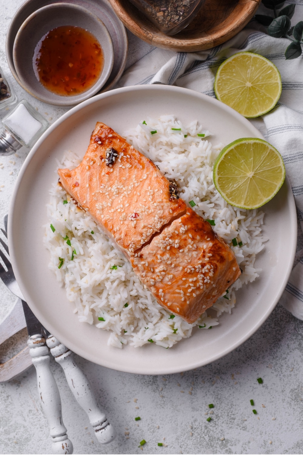 An overhead shot of a white plate with white rice and teriyaki salmon with sesame seeds on top. There is a halved lime next to the plate along with a fork and knife.