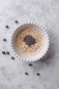 An overhead shot of a ceramic bowl filled with a milk mixture with chia seeds and oats on top. The bowl rests on a marble counter with a few scattered blueberries surrounding it.