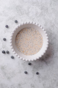 An overhead shot of a ceramic bowl filled with an oat, chia seed, and milk mixture on a marble counter with a few scattered blueberries surrounding it.