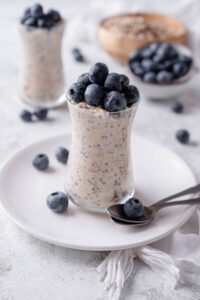 A glass jar filled with overnight oats and topped with fresh blueberries. The jar is on top of a ceramic plate with blueberries scattered around it, and a spoon in front.