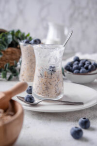 A half- eaten glass jar filled with overnight oats and topped with fresh blueberries. The jar is on top of a ceramic plate with blueberries scattered around it, and a spoon in front.