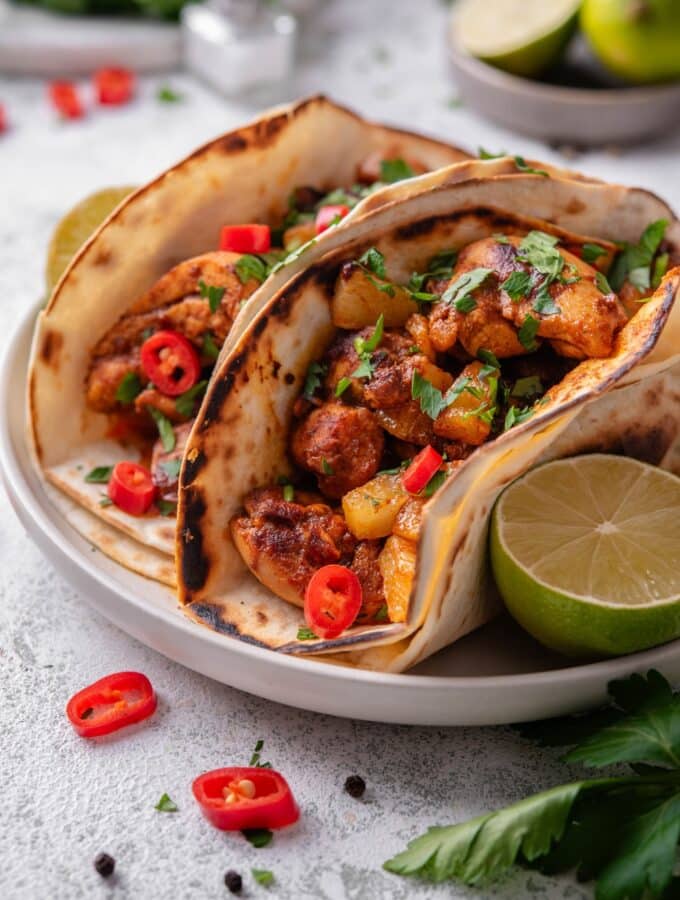 A plate with two chicken al pastor tacos with a halved lime on the side. The tacos contain chicken, pineapple, and peppers with cilantro on top. There is a bowl of limes in the background.