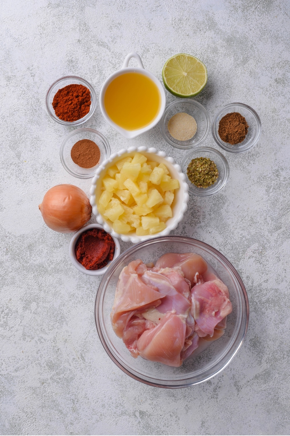 An overhead shot of several bowls in various sizes filled with ingredients for chicken al pastor including raw chicken thighs, pineapple chunks, pineapple juice, chili powder, and oregano. There is also an onion and halved lime.