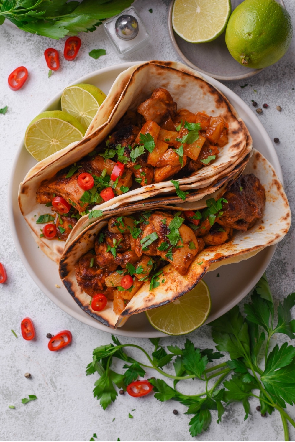 An overhead shot of a plate with two chicken al pastor tacos with halved limes on the side. The tacos contain chicken, pineapple, and peppers with cilantro on top. There is another bowl of limes on the side along with sprigs of cilantro sprinkled next to the plate.