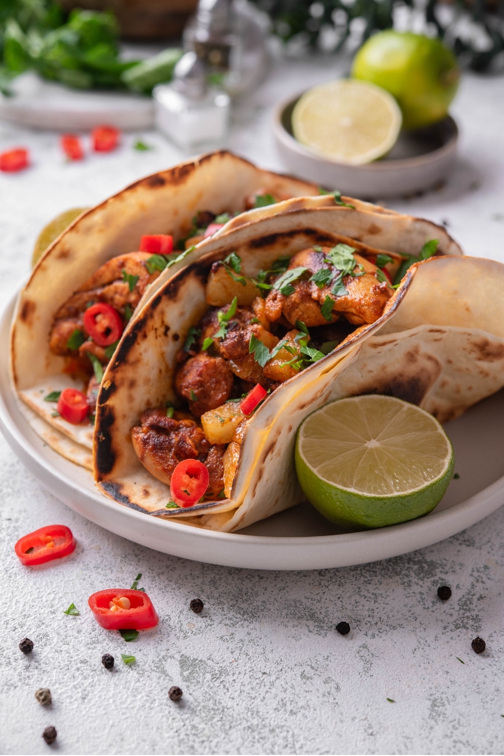 A plate with two chicken al pastor tacos with a halved lime on the side. The tacos contain chicken, pineapple, and peppers with cilantro on top. There is a bowl limes in the background.