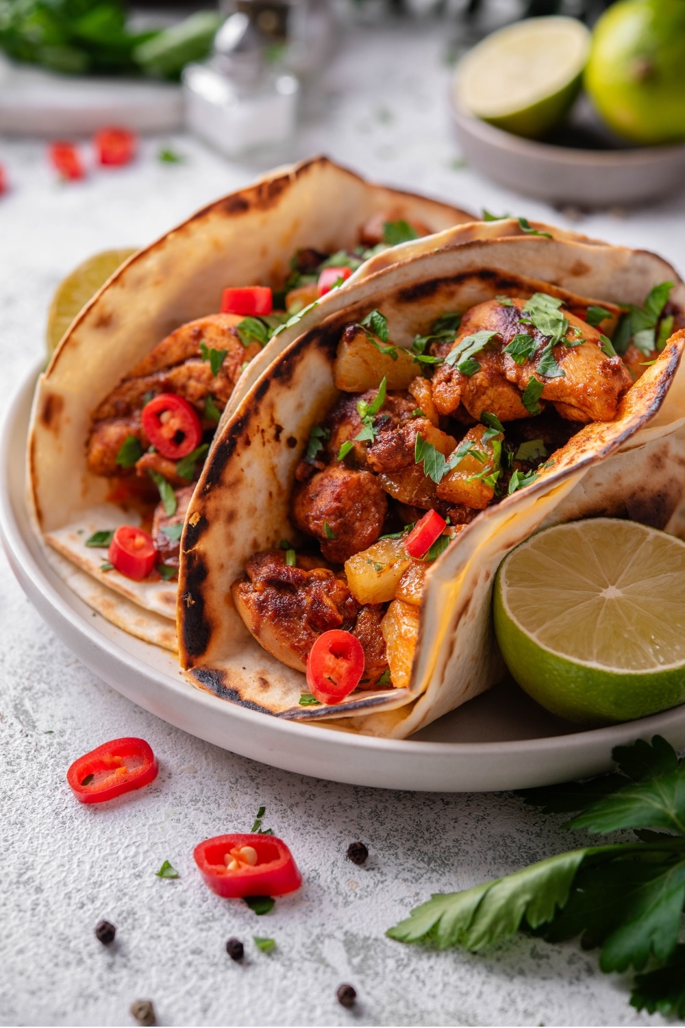 A plate with two chicken al pastor tacos with a halved lime on the side. The tacos contain chicken, pineapple, and peppers with cilantro on top. There is a bowl of limes in the background.