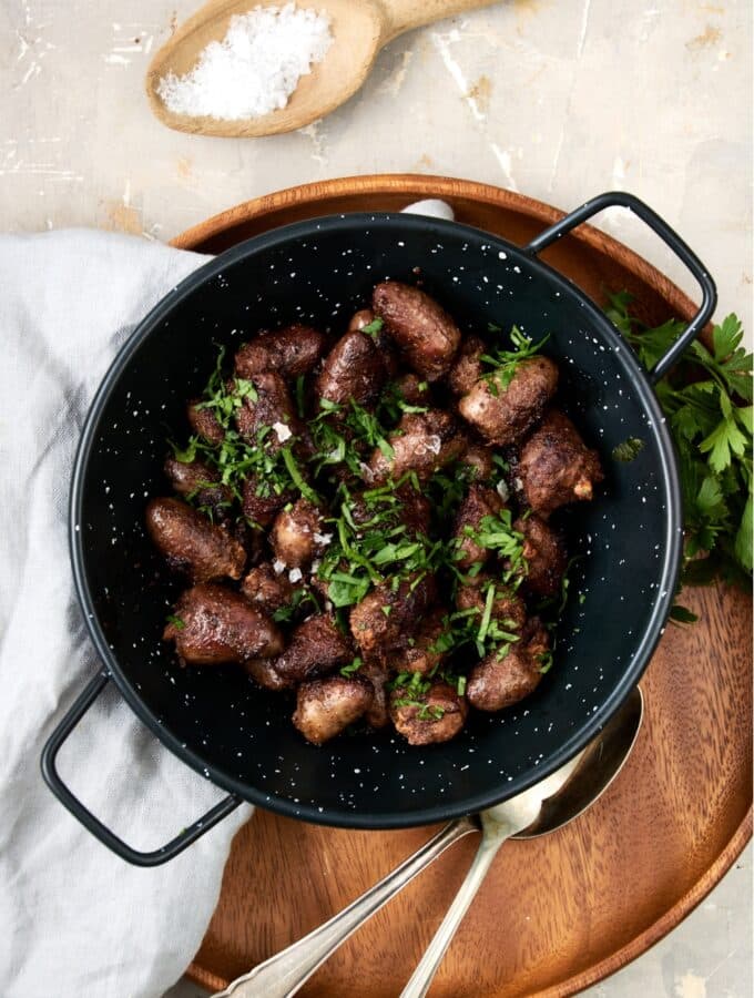 An overhead shot of a ceramic bowl filled with cooked chicken hearts topped with fresh parsley. The bowl is on a wooden server with a fork, spoon, and napkin next to the bowl.