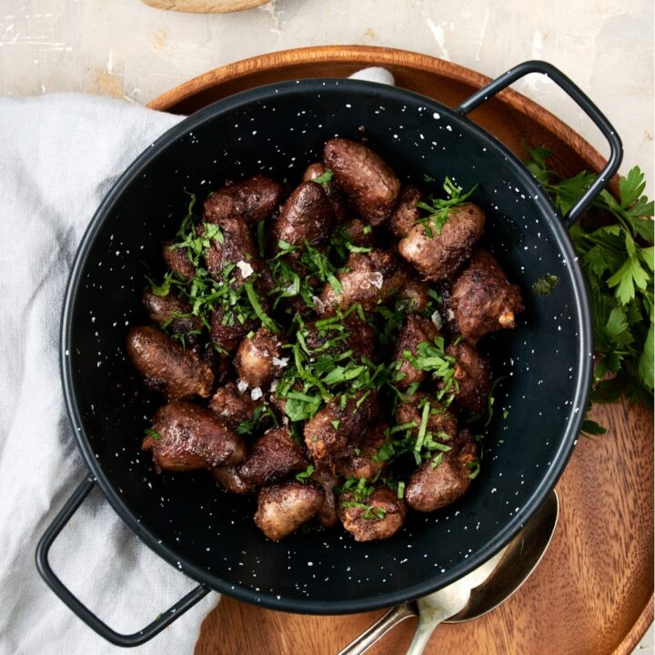 An overhead shot of a ceramic bowl filled with cooked chicken hearts topped with fresh parsley. The bowl is on a wooden server with a fork, spoon, and napkin next to the bowl.