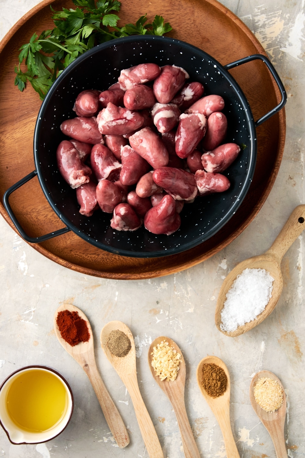 An overhead shot of chicken hearts ingredients. There are raw chicken hearts in a ceramic bowl on a wooden server, and several wooden spoons holding spices such as salt, smoked paprika, cumin, garlic powder, and onion powder.