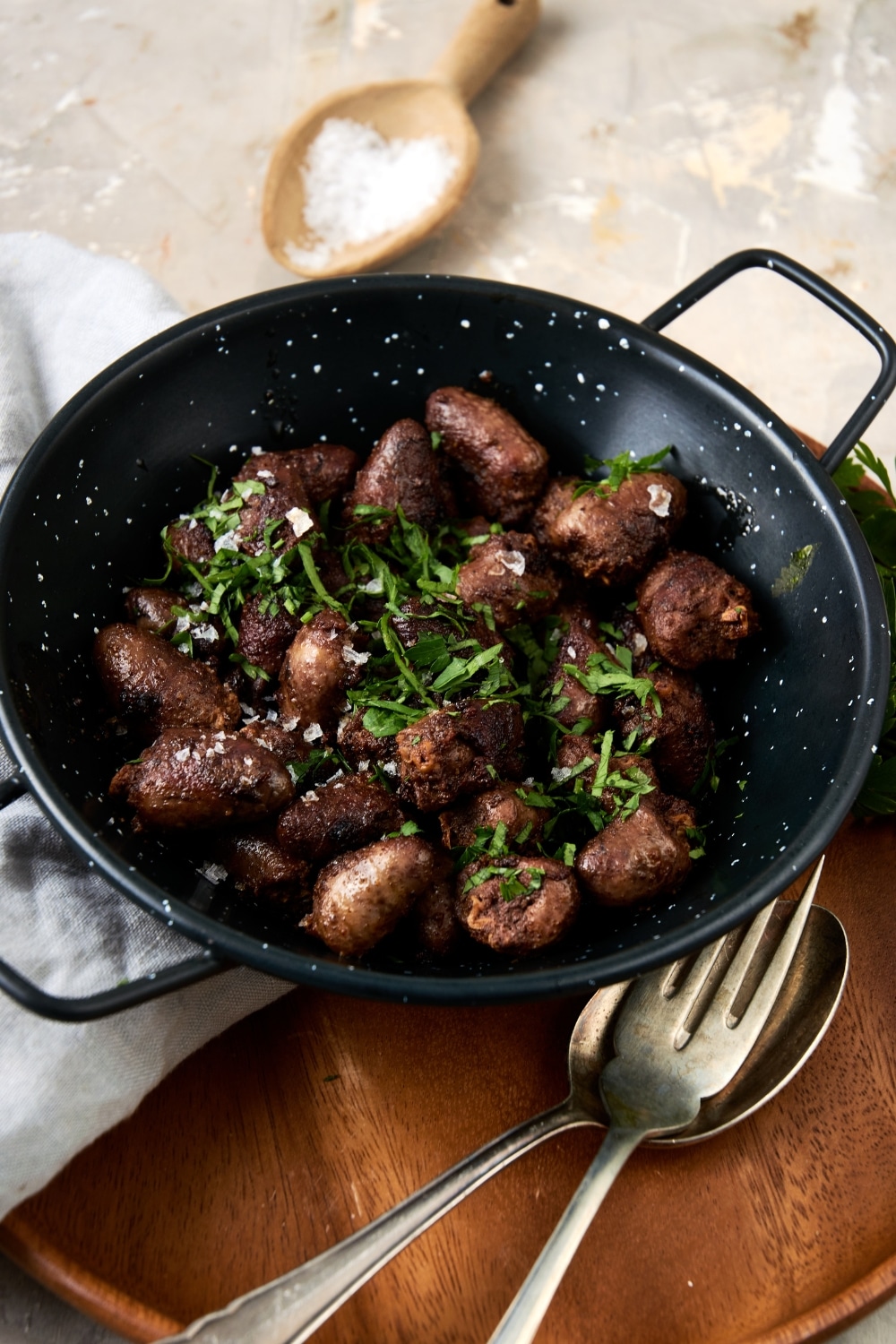 A ceramic bowl filled with cooked chicken hearts topped with fresh parsley. The bowl is on a wooden server with a fork, spoon, and napkin next to the bowl.