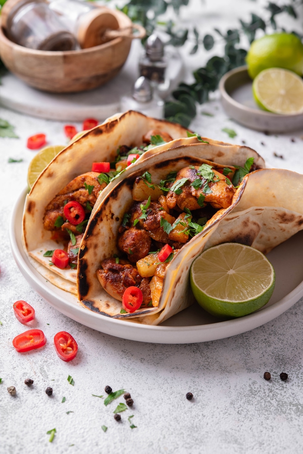 A plate with two chicken al pastor tacos with a halved lime on the side. The tacos contain chicken, pineapple, and peppers with cilantro on top. There is a wooden bowl with seasonings and limes in the background.