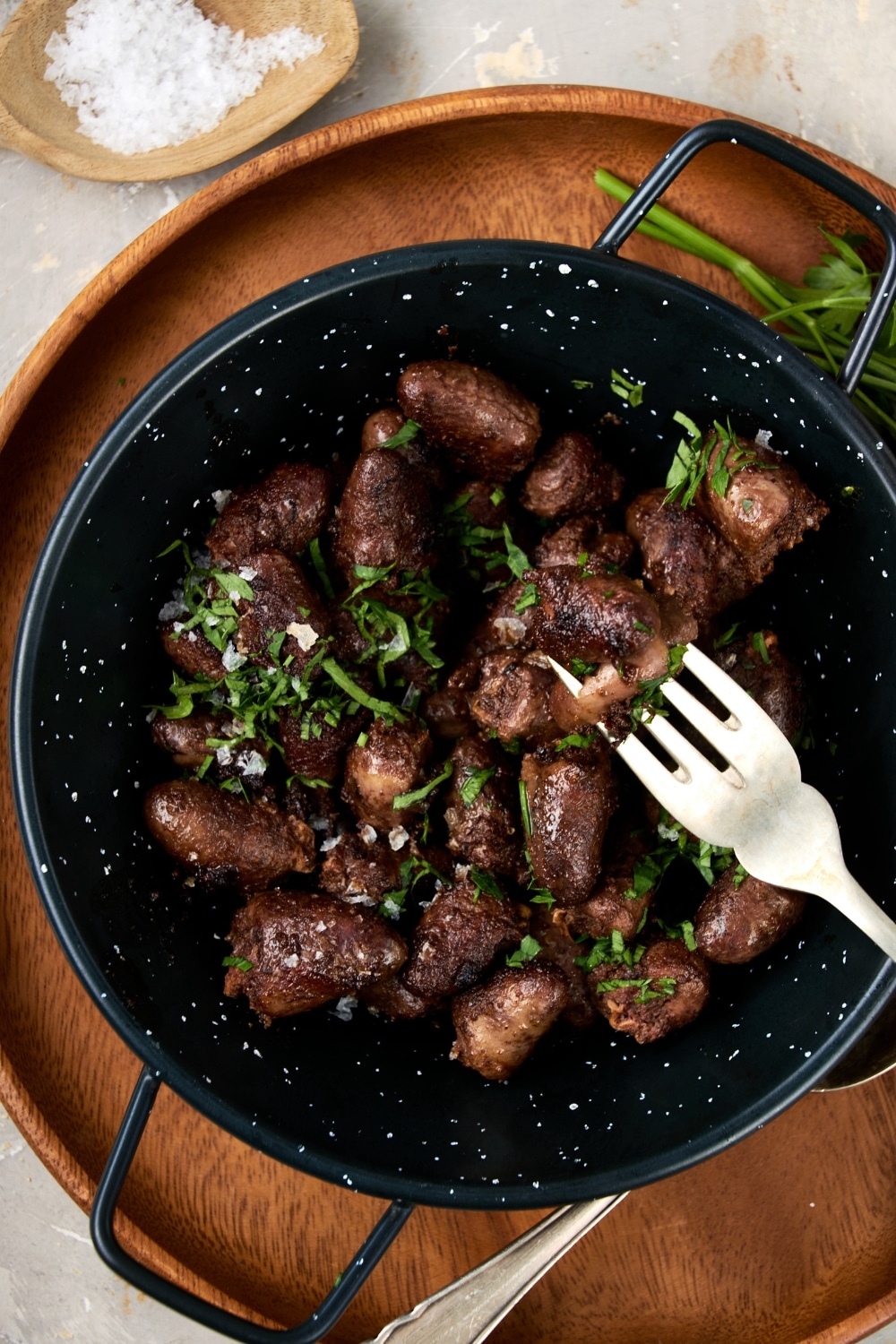 An overhead shot of a ceramic bowl filled with cooked chicken hearts topped with fresh parsley. The bowl is on a wooden server with a fork taking a bite of chicken hearts.