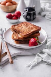 Four slices of french toast stacked on a white plate on top of a serving board. There is a strawberry and forks on the side.