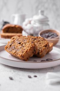 Three slices of low calorie zucchini bread with chocolate chips arranged on a white ceramic plate. There are a few chocolate chips scattered with another loaf of bread and bowl of chocolate chips in the background.