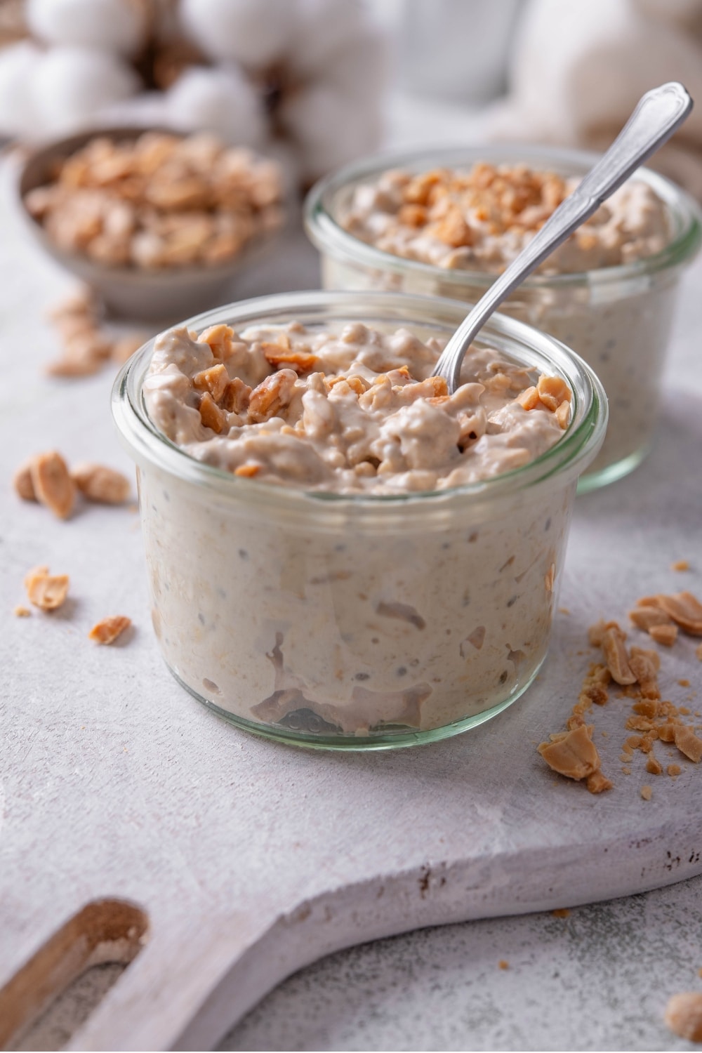 A small glass jar filled with peanut butter overnight oats on top of a wooden serving board with peanuts sprinkled around it. There is a spoon dipped into the jar.