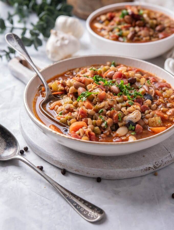 A bowl of bean soup in a ceramic soup bowl on a wooden serving board. There is a spoon dipped in the soup with another bowl of soup in the background.