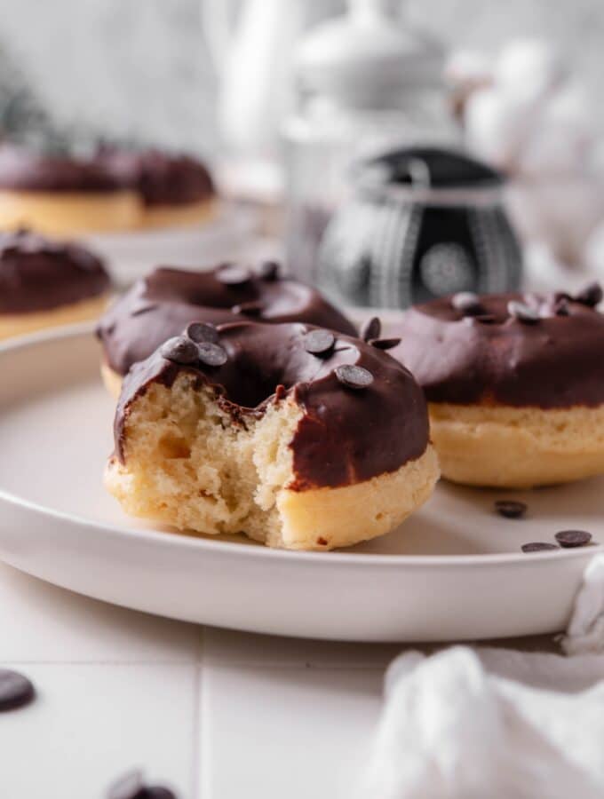 A ceramic plate with three baked donuts with chocolate frosting on top. The donuts are sprinkled with chocolate chips and one of the donuts has a bite taken out of it.