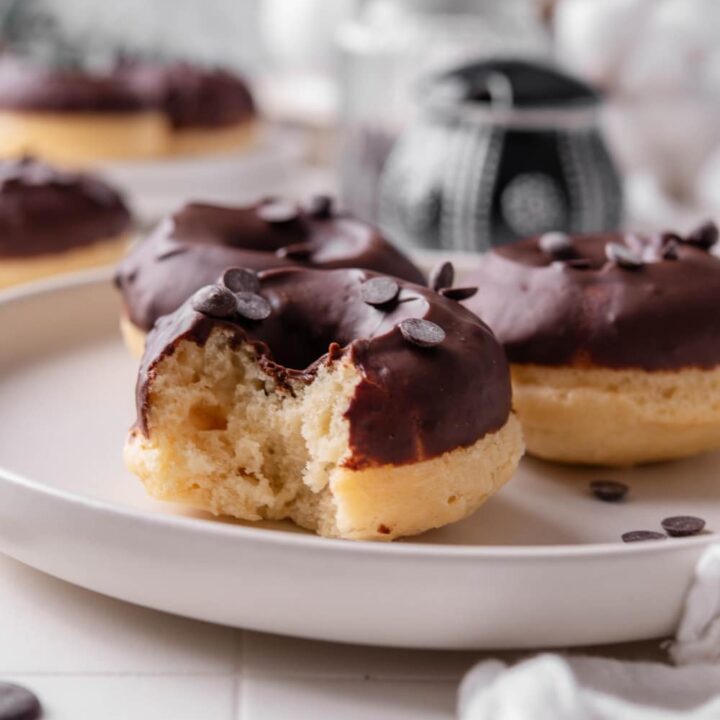 A ceramic plate with three baked donuts with chocolate frosting on top. The donuts are sprinkled with chocolate chips and one of the donuts has a bite taken out of it.