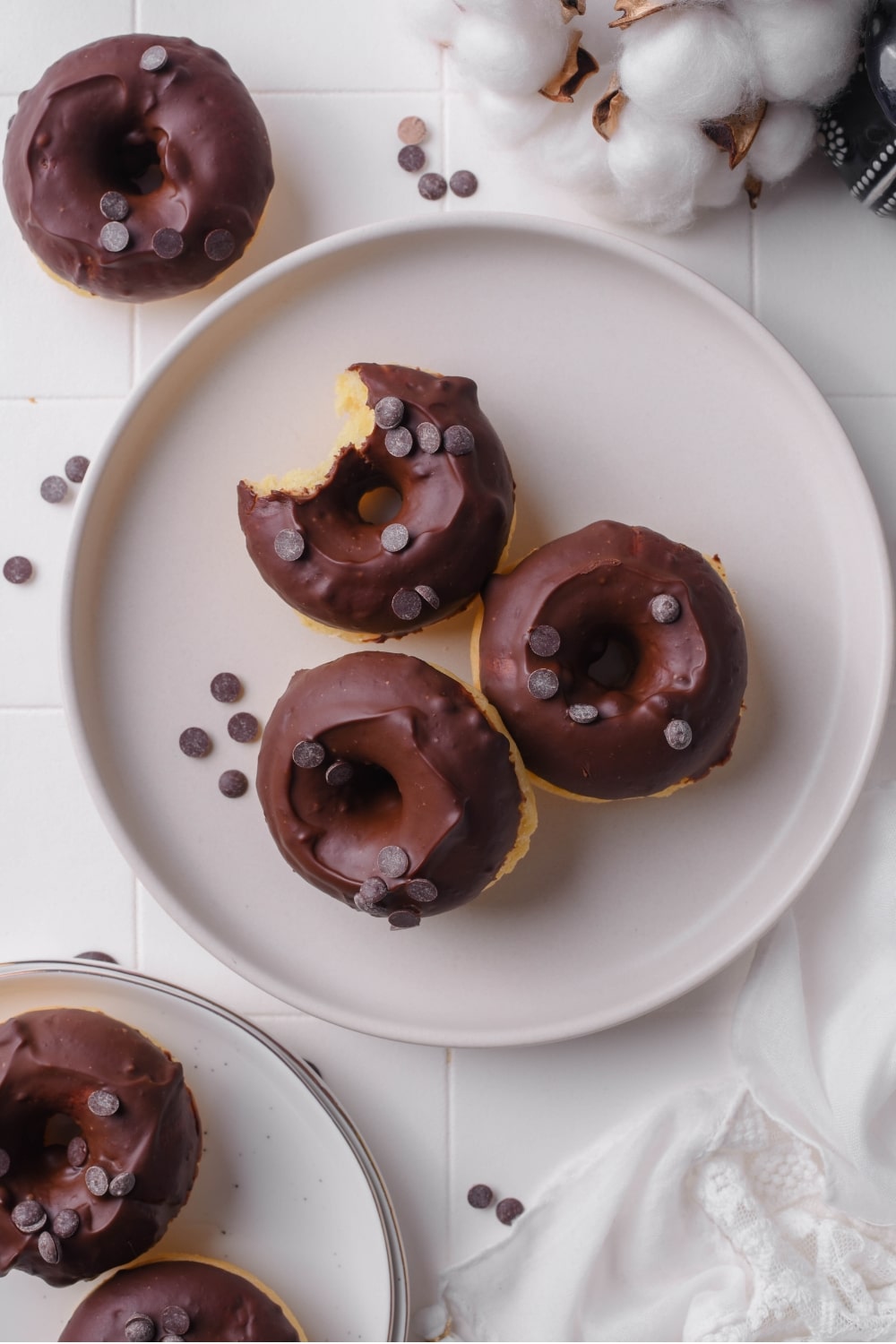 An overhead shot of three baked donuts on a ceramic plate. The donuts have chocolate frosting with chocolate chips sprinkled on top. There are additional donuts next to the plate.