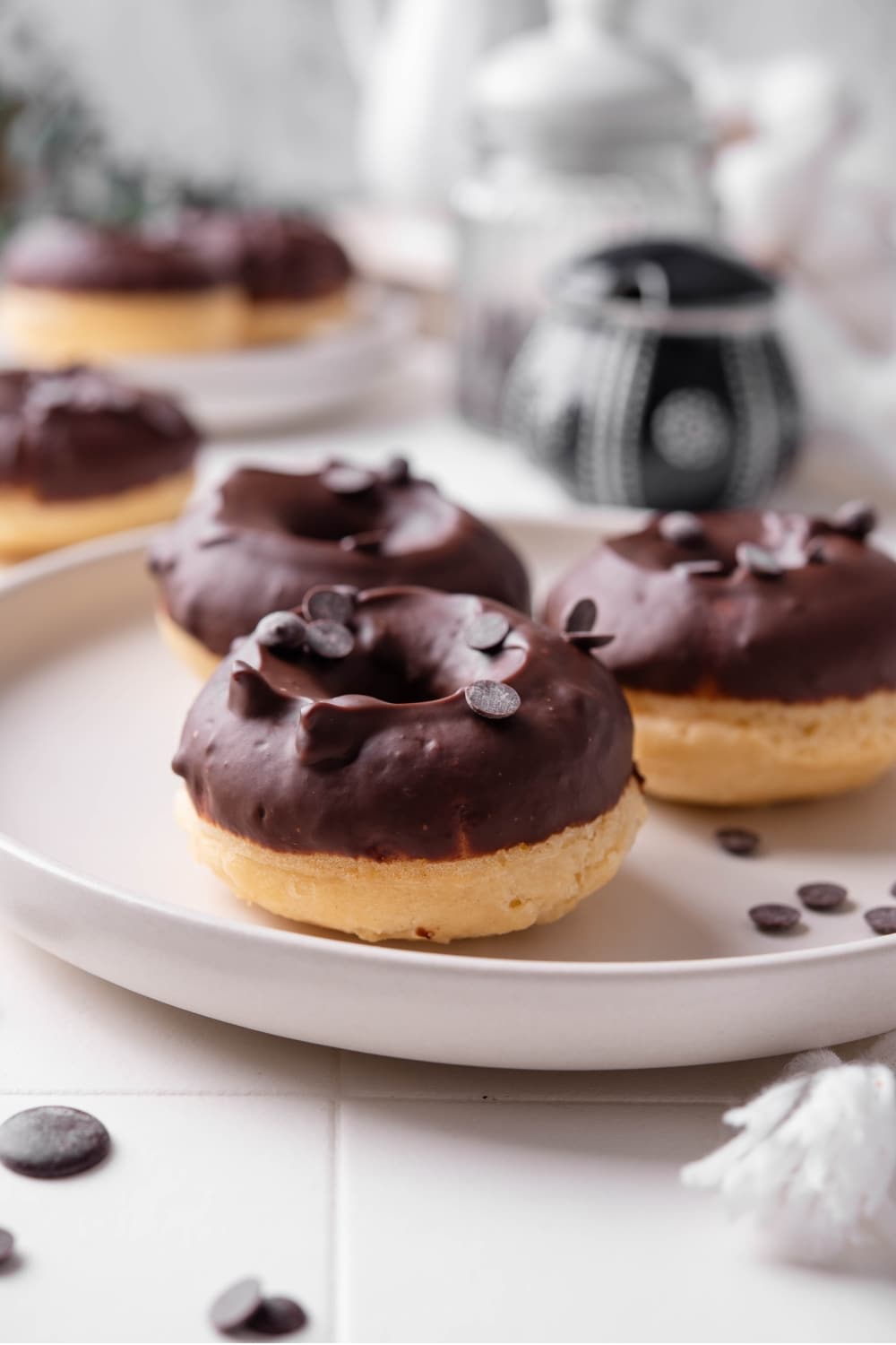 A ceramic plate with three baked donuts with chocolate frosting on top. The donuts are sprinkled with chocolate chips and there is an additional plate of donuts in the background.