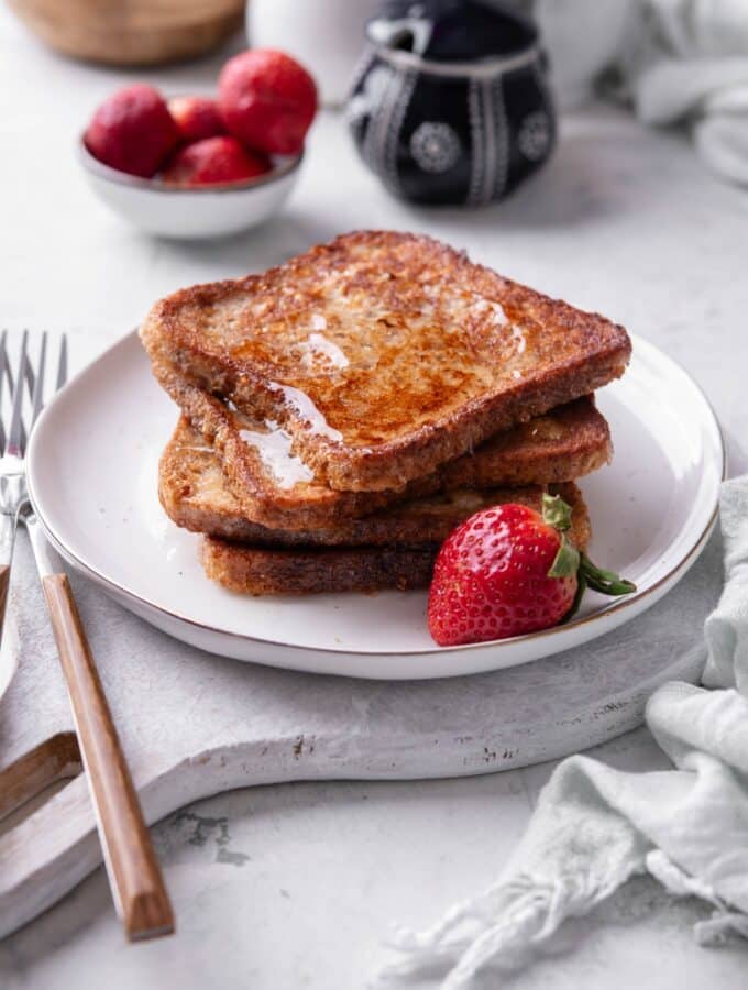 Four slices of french toast with syrup stacked on a white plate on top of a serving board. There is a strawberry and fork on the side.