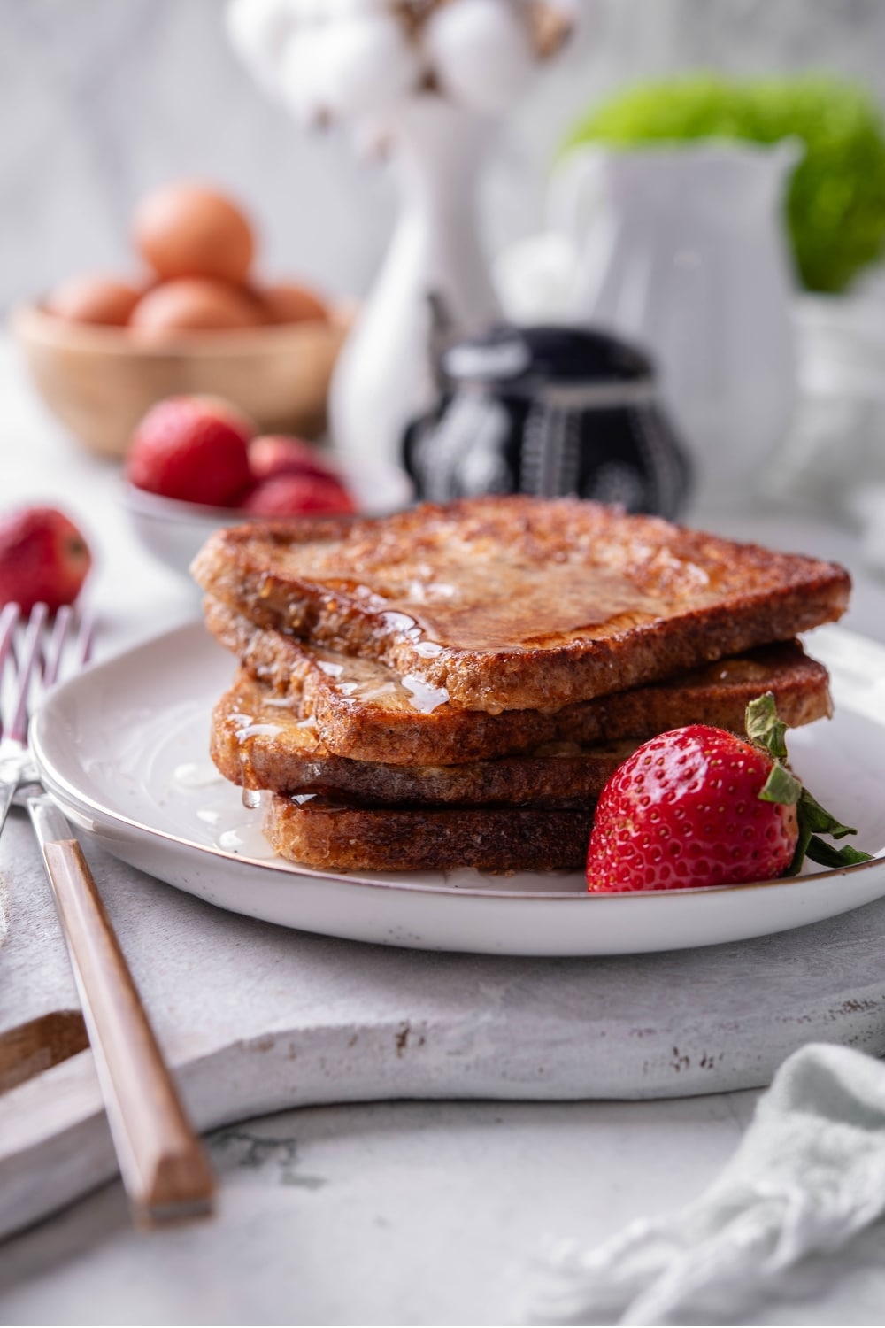 Four slices of french toast with syrup stacked on a white plate on top of a serving board. There is a strawberry and fork on the side.