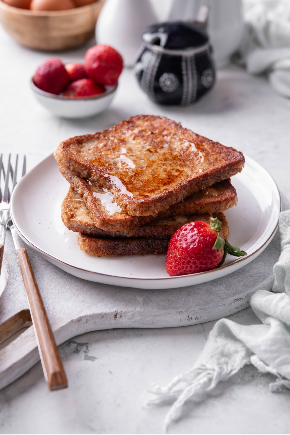 Four slices of french toast with syrup stacked on a white plate on top of a serving board. There is a strawberry and fork on the side.