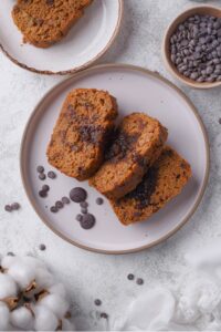 An overhead shot of three slices of zucchini bread with chocolate chips displayed on a ceramic plate. There are chocolate chips scattered on the plate and a small bowl of chocolate chips next to the plate.