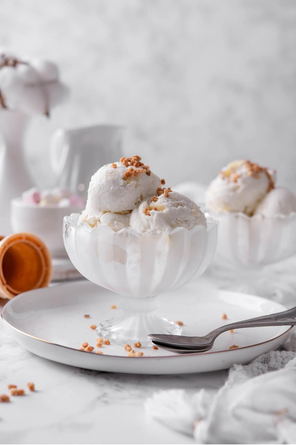 Three scoops of vanilla protein ice cream in a glass sundae bowl on a white plate with a spoon to the side. There are some nuts sprinkled on top.