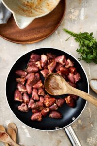 An overhead shot of a skillet with chicken hearts cooking in spices. There is a wooden spoon resting in the skillet.