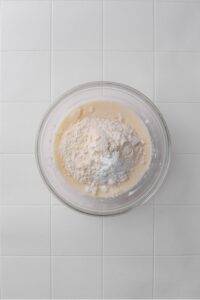 A glass bowl filled with a whisked coconut milk mixture with flour and baking powder on top.