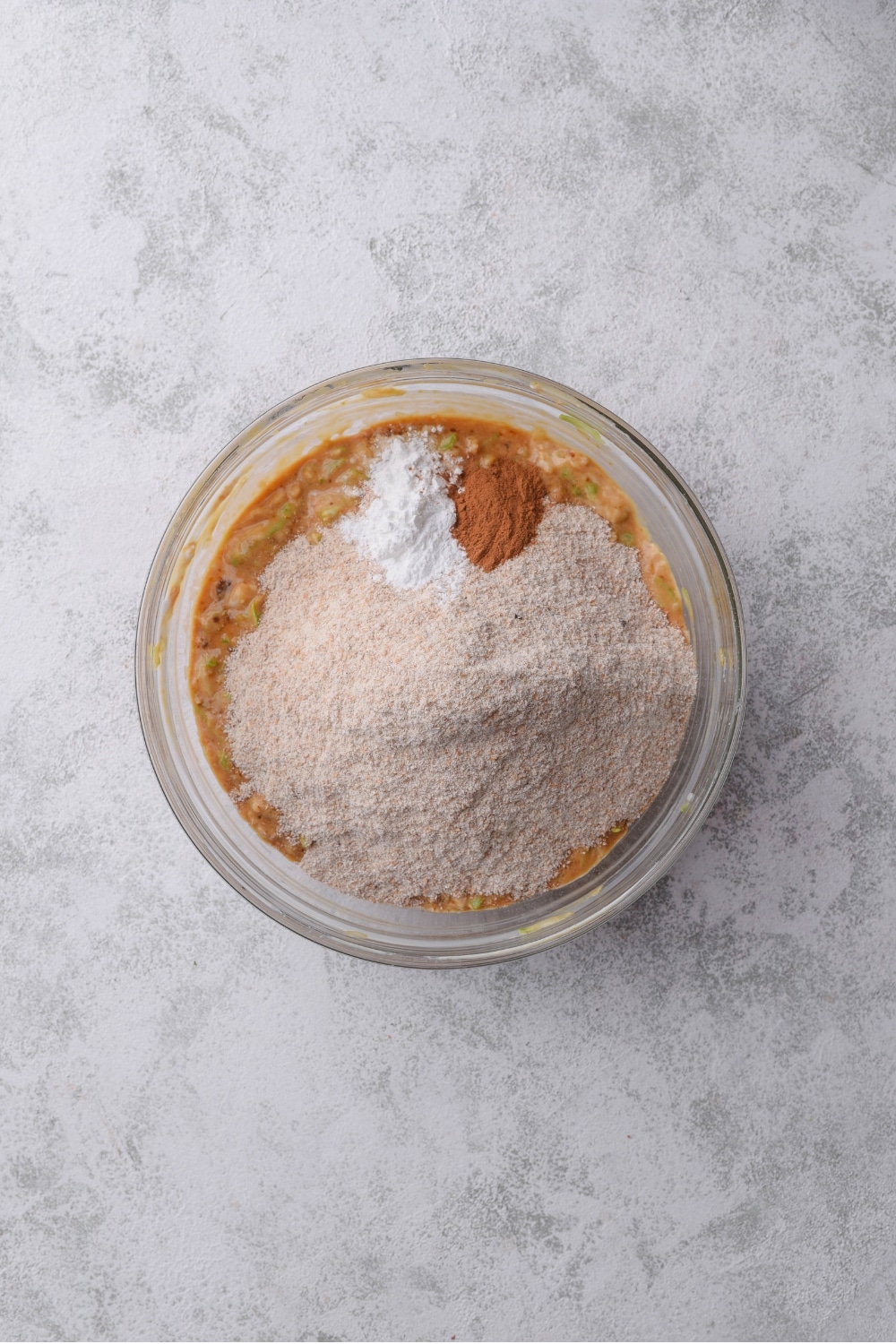 A glass bowl filled with zucchini bread batter with flour, cinnamon, and baking soda on top.