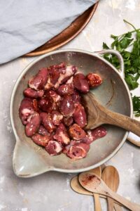 A ceramic mixing bowl filled with raw chicken hearts, oil, and seasonings being stirred with a wooden spoon.