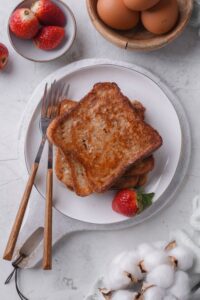 An overhead shot of four slices of french toast with syrup stacked on a white plate. The plate is on top of a serving board with a strawberry and fork on the side.