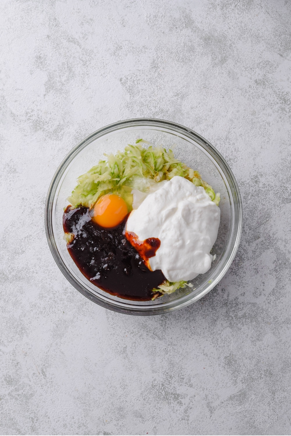 A glass bowl filled with shredded zucchini, raw egg, greek yogurt, vanilla, and syrup.