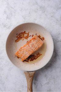 A ceramic skillet with salmon cooking in olive oil topped with sesame seeds.