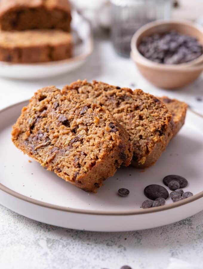 Three slices of low calorie zucchini bread with chocolate chips arranged on a white ceramic plate. There are a few chocolate chips scattered with another loaf of bread and bowl of chocolate chips in the background.