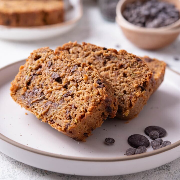 Three slices of low calorie zucchini bread with chocolate chips arranged on a white ceramic plate. There are a few chocolate chips scattered with another loaf of bread and bowl of chocolate chips in the background.